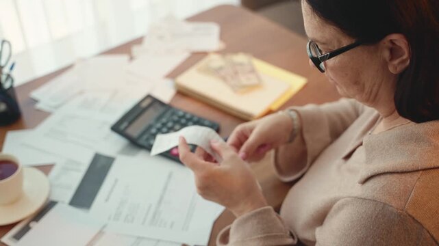 Elderly woman stressed pensioner woman is looking through receipts for utility bills sitting at desk planning budget. Mature lady checking taxes bank account balance. Expenses medical insurance policy