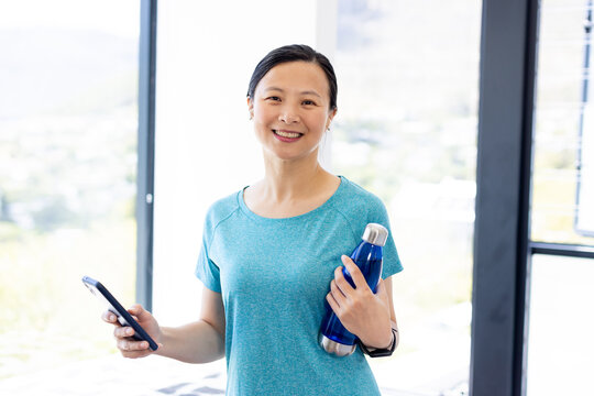 Asian woman smiling, holding phone, blue bottle, wearing teal shirt and fitness watch in home gym