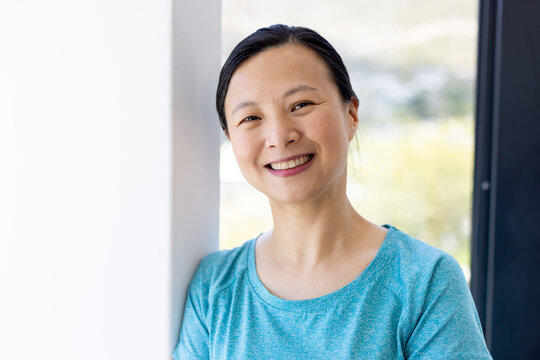 Asian woman leaning against white wall by large window, smiling and facing camera in teal top
