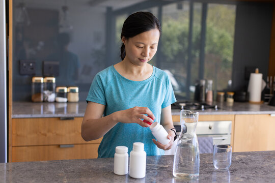 Asian woman standing at stone kitchen island, removing red cap from pill bottle, holding carafe