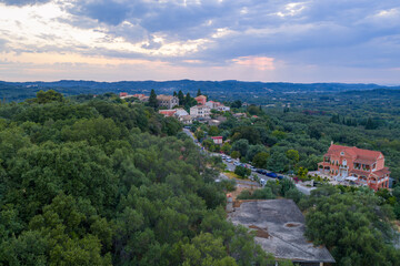 Fototapeta premium Aerial drone view of a traditional Mediterranean village in Corfu, Greece, with colorful houses, stone rooftops, winding roads, lush green hills and surrounding countryside landscape under summer sky.