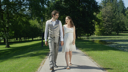 Fototapeta premium Young pregnant couple strolling in park deeply gaze at each other holding hands