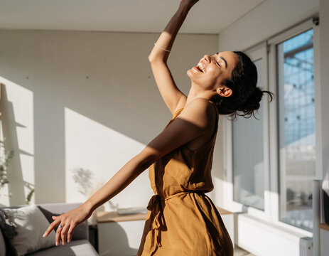 Woman Joyfully Dancing Alone in Her Apartment. Levity & Laughter / Joyful Micro Moments. sunny interior photograph of a young woman dancing with eyes closed in her bright apartment living room.
