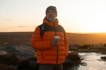 Man stands on a rocky hill with coffee during sunset in nature © Iryna