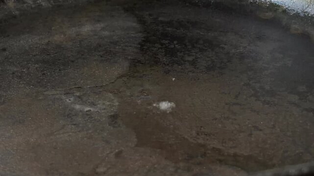 A single drop of water falls onto the dark, heated surface of a frying pan. Upon contact, the liquid instantly vaporizes, creating a burst of white steam and a sharp, aggressive sizzle.