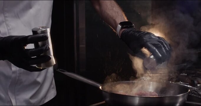 Chef adds spices to meat fillet on hot frying pan in kitchen closeup. Master uses seasoning for better taste of dish with veal in fancy restaurant. Dinner preparation