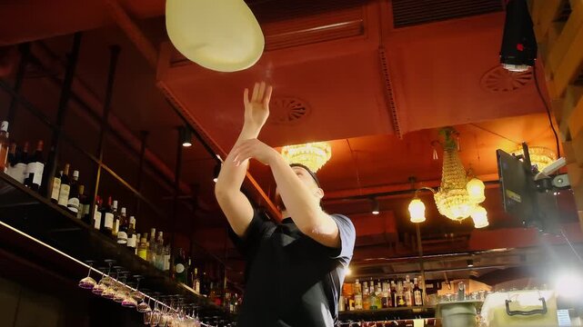 Man pizzaiolo confidently tosses pizza dough into air forming thin round base. Male throws dough upward and lets spin gently. Slow motion