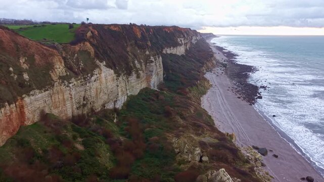 White chalk cliffs and eroded coastal slope of Plage de Saint-Jouin-Bruneval stretch above a long pebble beach and grey English Channel surf in Normandy, France, drone slow gliding.