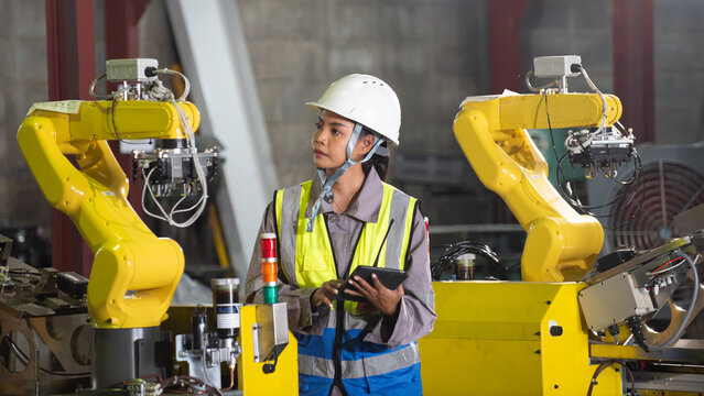 Industrial plants factory technician robot automation safety helmet tablet inspection in industrial floor as focused worker guides yellow robotic arm inside modern production area