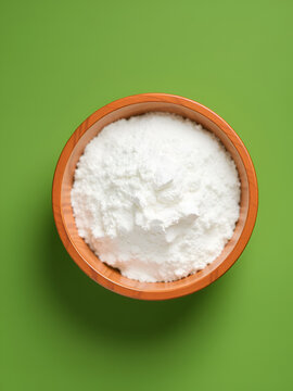 Food additive E575 in wooden bowl on green, top view. Glucono Delta-Lactone (GDL) or Gluconolactone used as sequestrant, acidifier, leavening agent. White powder, oxidized derivative of glucose