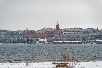 Obraz premium Historic red brick building surrounded by snowy trees near a calm body of water in Flensburg