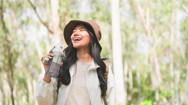 Cheerful young Asian woman with a backpack and bucket hat laughing and holding a reusable water bottle during a hike in a lush green forest.