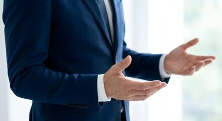 Businessman in a dark blue suit gesturing with open hands during a presentation or speech in an office setting