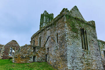 Overcast sky above the historic Timoleague Abbey ruins in County Cork, Ireland. Ancient stone walls, covered in vibrant green moss, feature arched windows.