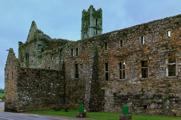 Ancient stone ruins of a historical Irish abbey feature crumbling walls, an imposing tower, and open windows. Green moss covers the decaying structure.