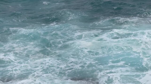 Ocean waves crash and froth in turbulent motion on a rocky shoreline from an aerial viewpoint.