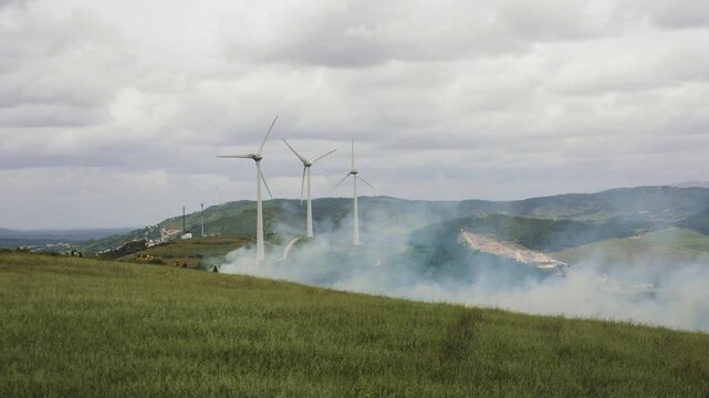 Daytime 4K timelapse of wildfire smoke and firefighters with tanker trucks. Wind turbines stand in background, symbolizing climate and energy transition