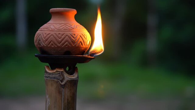 Close up of a handcrafted terracotta oil lamp with a warm glowing flame on a bamboo pole at dusk in a lush green forest setting with blurred background and soft natural light illuminating the