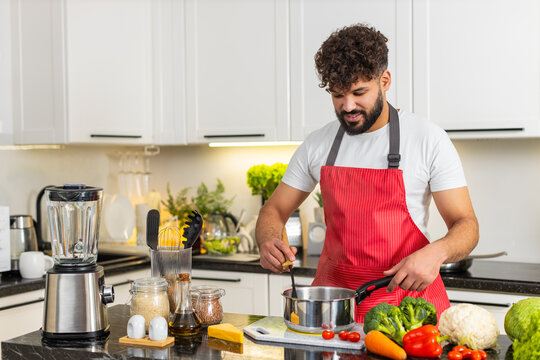 Arabian man stirring warm soup in pot on stove in kitchen cooking hearty lunch for healthy nutrition. Young guy focused on meal prep enjoying easy recipe for family dinner in apartment home indoors.