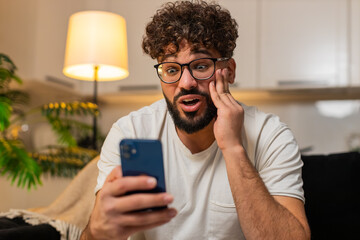 Arabian man at home checks smartphone reads happy message reacts with wow covers mouth smiles....