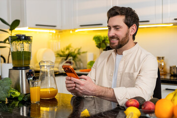 Middle-aged man smiling while typing on smartphone in home kitchen beside fresh fruit juice and...