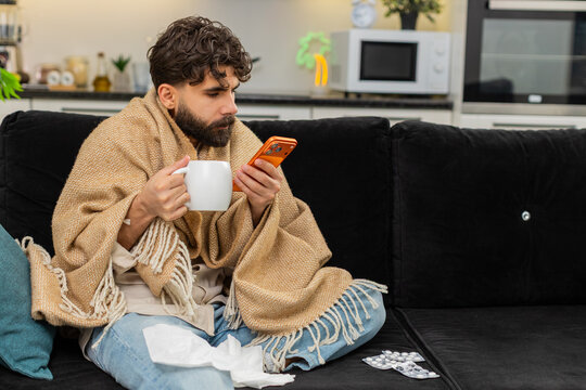 Indian man at home wrapped in blanket looks at smartphone reading health tips from doctor feels weak. Arabian guy on sofa holds tissue by nose cup of tea and pills near hand checks mobile phone sadly.