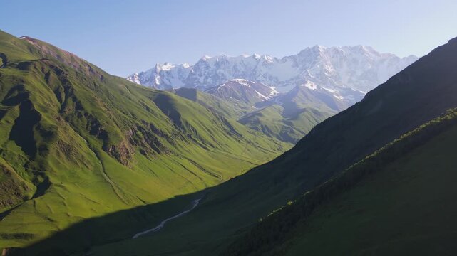 Aerial drone view moves forward in Svaneti between Mestia and Ushguli, a U shaped green valley, braided stream, long shadows, rocky outcrops, glacial cirques, snow clad peaks