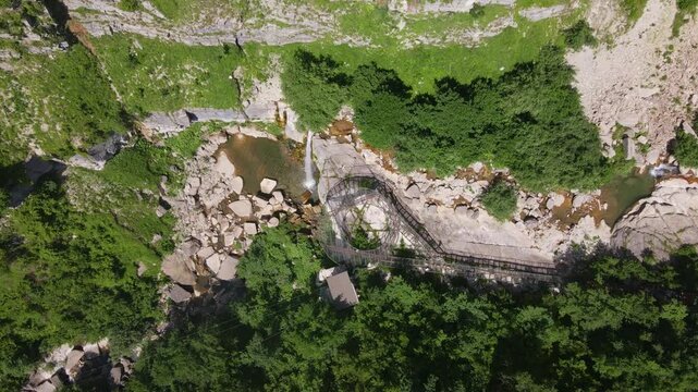 Aerial view of a spiral observation bridge over a rocky stream and waterfall in Svaneti, Georgia. Rushing water moves as tiny figures walk in bright midday light.