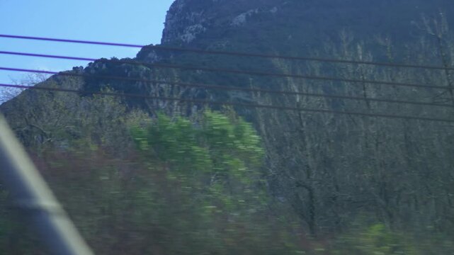 A panoramic view of forested mountains and rocky peaks from a train window in Italy. Dense green forests conceal the mountain slopes beneath a bright sky.