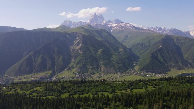 Aerial shot descends over Svaneti in Georgia toward Ushba, showing conifer forest, villages, fields, narrow roads, and terraces under clear daylight with wide framing
