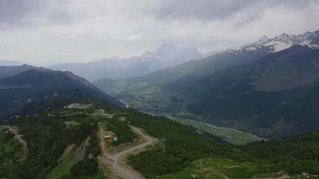 Aerial drone shot moves along a green ridgeline with a zigzag dirt road, ski lift pylons, and buildings, revealing a river valley near Mestia and Ushguli in Upper Svaneti, Georgia.