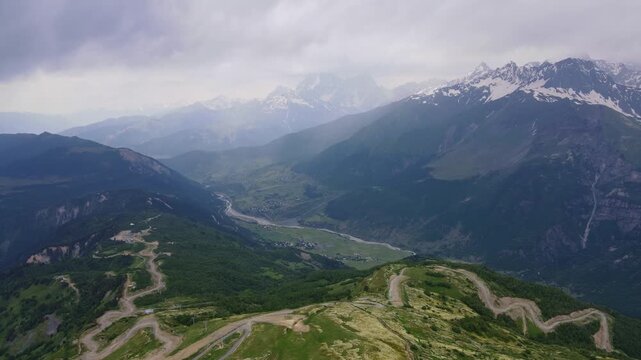 Aerial view over a high Svaneti ridge in Georgia shows a serpentine switchback road, valley villages, a meandering river, forested slopes, and snow capped Caucasus peaks.