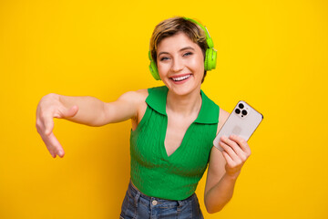 Naklejka premium Happy young woman with green headphones smiles and reaches toward the viewer while holding a phone against a bright yellow background