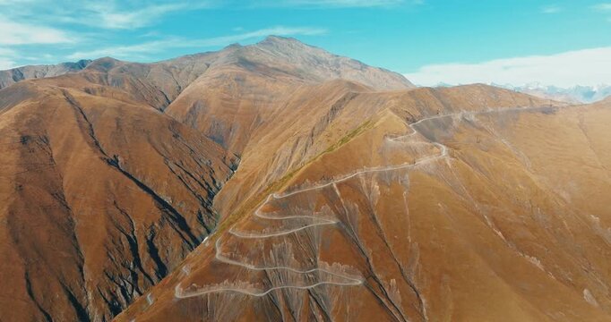 Aerial drone shot of the winding Abano Pass road in Tusheti, Georgia, featuring a slow zoom-in towards the majestic mountain peaks under a clear blue sky.