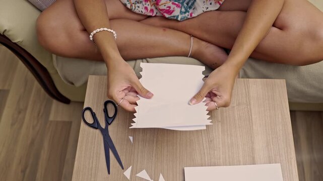 Showcasing the meticulous process of cutting white paper with scissors, creating intricate zig-zag patterns for a handmade craft project. An engaging and creative DIY activity on a wooden table.