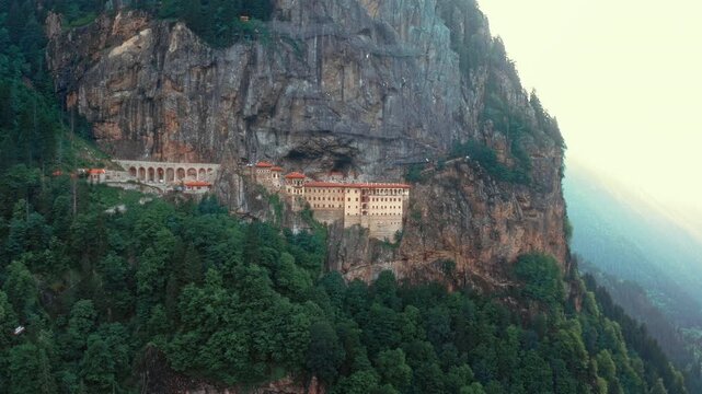 Aerial drone shot of Sumela Monastery in Trabzon with beautiful sun flare. Historic monastery on cliffs surrounded by misty forests at sunset.