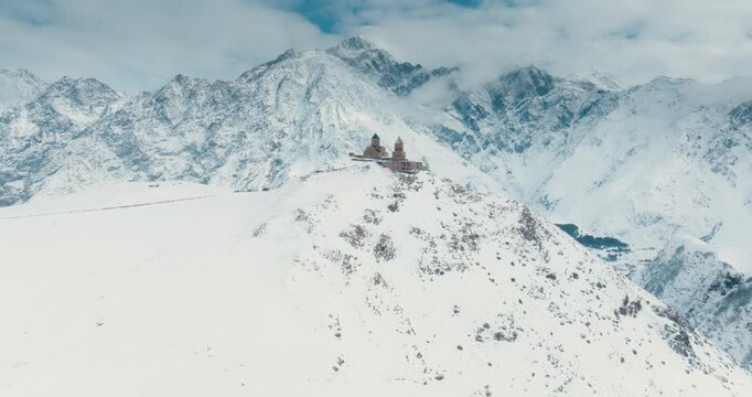 Drone zoom in shot of Gergeti Trinity Church against snowy Caucasus Mountains in Kazbegi, Georgia. Majestic winter landscape and historic architecture.