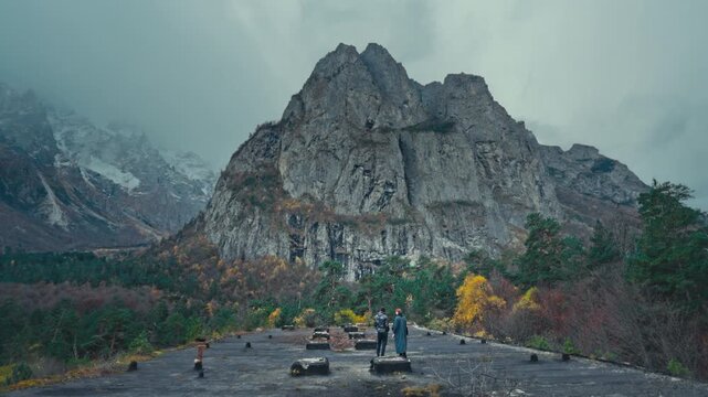Wide aerial shot of two people overlooking a vast mountain valley and massive rock formations under a misty, dramatic sky in North Ossetia.