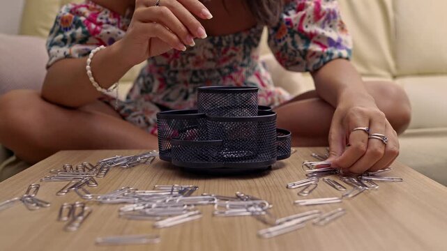 Close-up of a woman's hands meticulously organizing silver paper clips into a black mesh desk organizer. Demonstrates focus and productivity in a tidy home office environment.