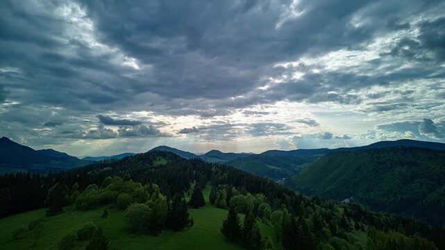 Aerial drone hyperlapse of moving clouds and sunbeams over mountain forest and meadows.