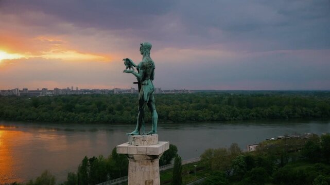 Aerial drone orbit of the Victor (Pobednik) monument in Belgrade at sunset. The bronze statue overlooks the confluence of the Sava and Danube rivers during a dramatic golden hour.