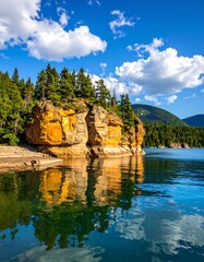 Sunny day shot shows rocky outcropping and trees mirrored in a blue lake with a bright, cloudy sky above