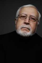 Obraz premium Older man with glasses and white beard poses against dark background, showing a serious expression with direct gaze