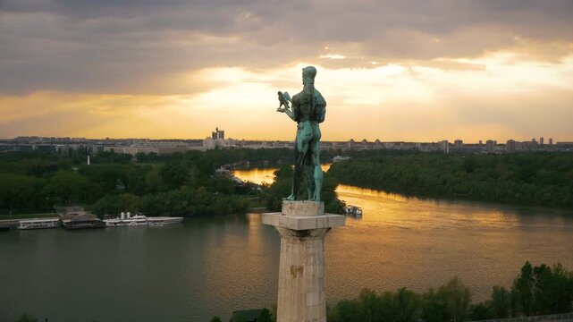 Cinematic drone shot of the Victor (Pobednik) monument in Belgrade during a golden sunset. The iconic statue stands tall over the confluence of the Sava and Danube rivers.