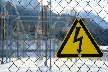 Triangular yellow high-voltage danger sign on fence, warning for electric shock, power transformer station out of focus behind, electrical safety symbol, utility infrastructure