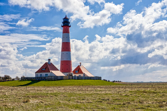 The red and white striped Westerheversand lighthouse in Schleswig-Holstein, Germany, surrounded by vast green salt marshes under a partly cloudy sky