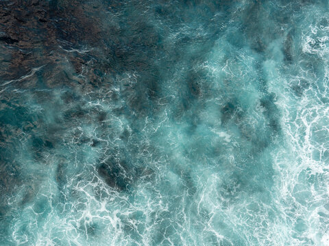 A top-down aerial view captures turquoise ocean waves swirling over dark rocky seabed in a dynamic natural pattern.