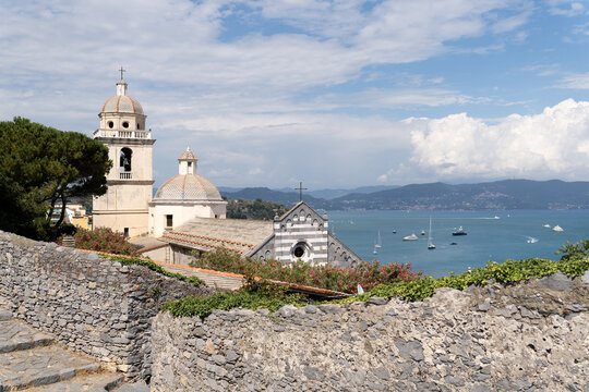 A historic seaside church with domed rooftops overlooks a calm blue bay dotted with sailboats under a bright sky.