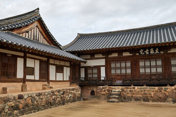 A serene eye-level view of a traditional Korean Hanok house courtyard in Andong Hahoe Village, South Korea, featuring weathered wood, stone foundations, and dark tiled roofs under a cloudy sky