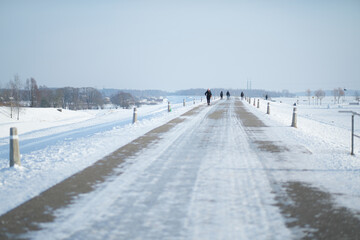 A long snowy road stretches through a bright winter landscape as people walk in the distance, creating a serene scene of cold weather, open space, and quiet seasonal atmosphere.